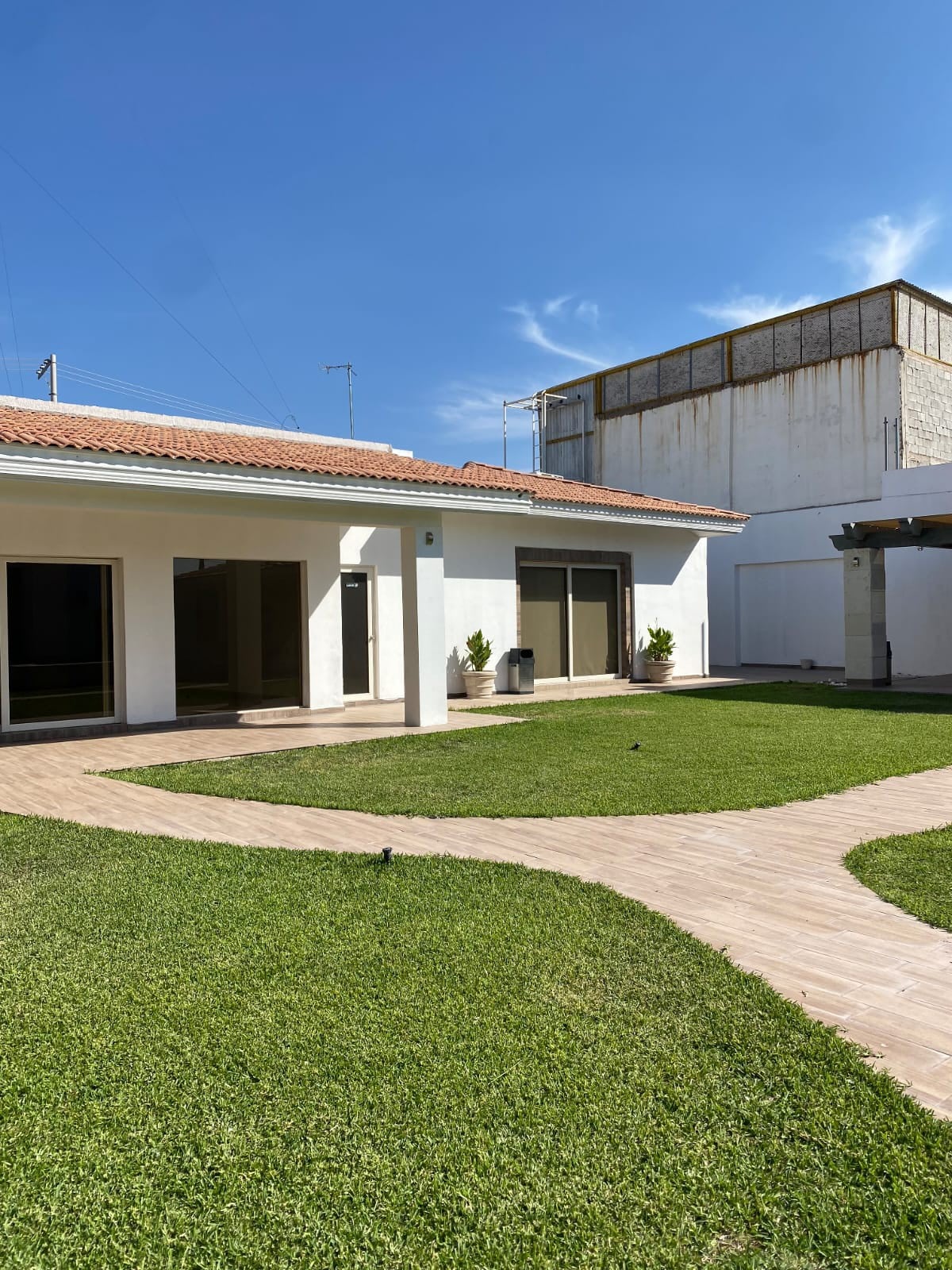 The outdoor area features a well-maintained lawn with pathways leading to various sections of the property. A covered patio is visible, alongside modern glass doors that connect the interior spaces to the serene surroundings, under a clear blue sky.