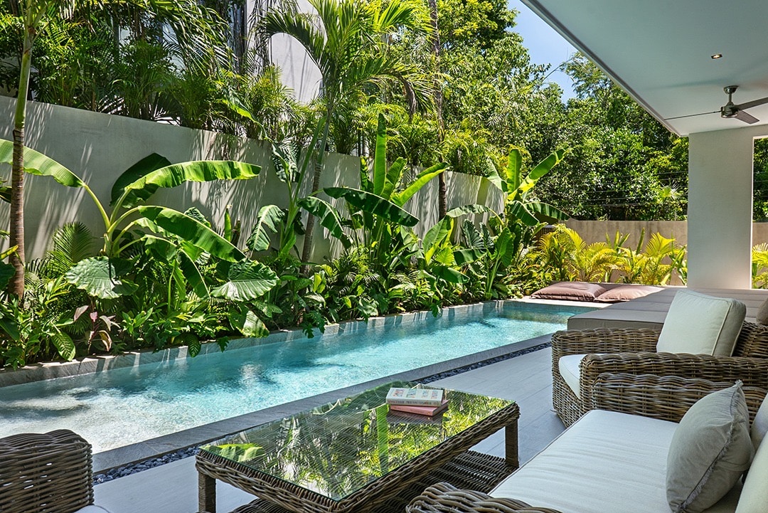 A serene outdoor area is showcased with a clear pool bordered by lush green plants. Comfortable seating is provided with woven furniture, positioned nearby a sleek glass table. Sunlight filters through the foliage, creating a peaceful retreat.