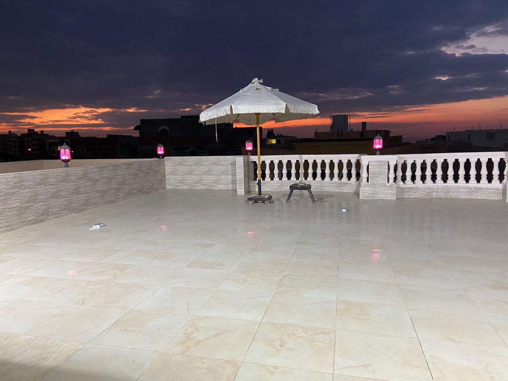 A spacious rooftop area features a tiled floor with ample open space for relaxation. A white umbrella provides shade, while lanterns hang along the edges, casting a soft glow. The backdrop showcases a colorful sunset sky, creating a serene ambience.