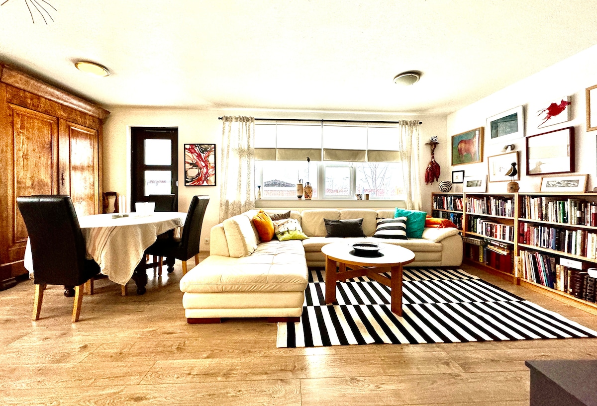 An inviting living room features a spacious sectional sofa with colorful cushions, a round coffee table, and a striped rug. A dining table sits in the corner, while bookshelves line one wall, enhancing the cozy atmosphere. Large windows allow natural light to fill the space.