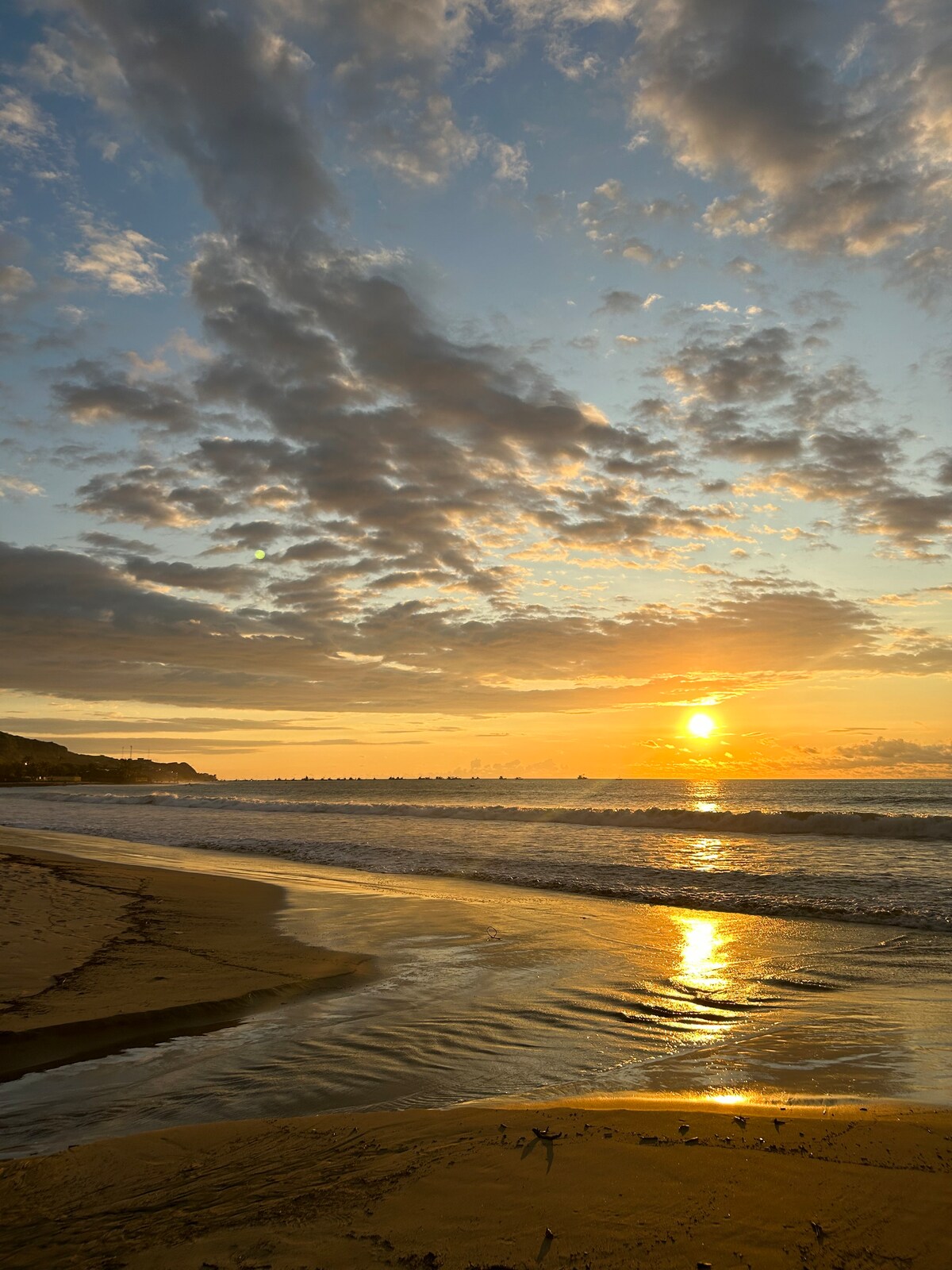 A tranquil beach scene is captured at sunset, where golden light reflects off gentle waves. Soft sand stretches along the shore, with patterns created by the receding tide. Clouds are scattered across the sky, adding to the serene atmosphere of the coastal setting.
