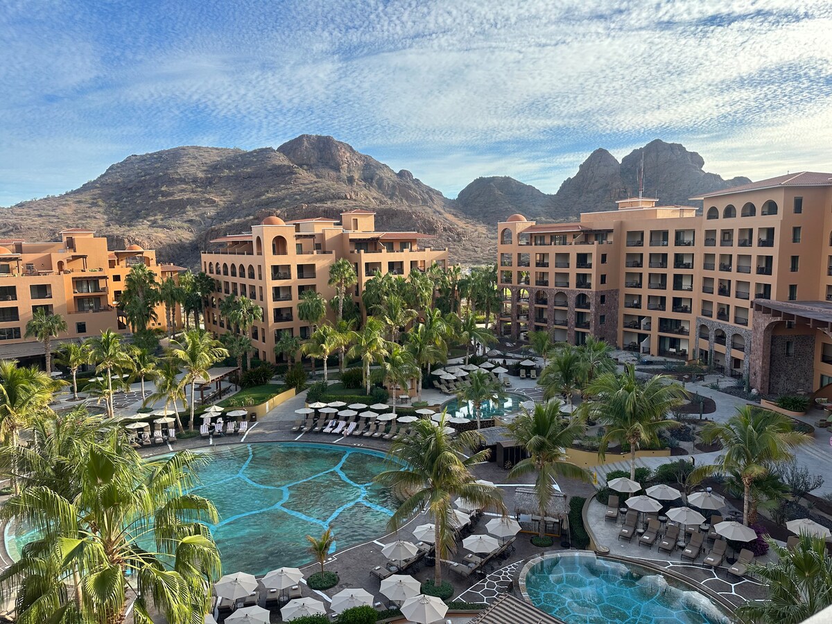 The expansive resort complex features a series of terracotta buildings surrounded by lush palm trees and inviting swimming pools. The backdrop includes rugged mountains under a vibrant sky, creating a serene atmosphere that complements the resort's architecture.