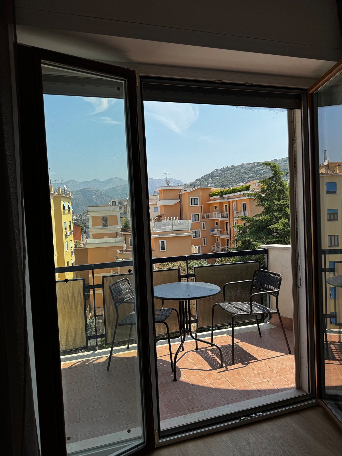 A view from the penthouse showcasing a balcony with two chairs and a small table. The surrounding buildings in varying colors complement the mountains in the distance, with natural light illuminating the area through the open glass doors.