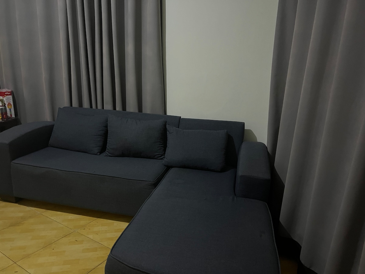 A modern sectional sofa in a muted tone occupies the corner of the room. Soft cushions enhance comfort, while flowing gray curtains drape elegantly over the large window, contributing to a calm and relaxed atmosphere.
