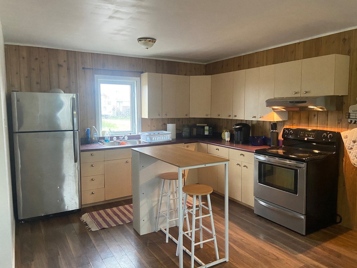 A functional kitchen space is shown, equipped with modern appliances including a stainless steel refrigerator and an oven. Light wooden cabinetry complements the warm-toned walls. A central island with two simple stools provides additional seating. Natural light enters through a window, illuminating the room.