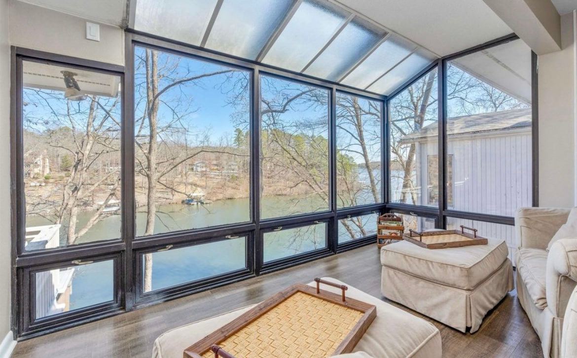 A glass-enclosed sunroom offers panoramic views of the lake and surrounding trees. Comfortable seating is positioned to enjoy the natural light and serene scenery. A pair of wooden trays sits on the soft ottomans, adding convenience to the relaxing space.
