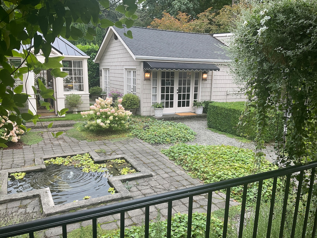 The image captures a serene outdoor garden space, featuring a small pond surrounded by greenery and cobblestone pathways. A cottage structure is visible in the background, framed by lush plants and hydrangea blooms, all under a softly lit sky.
