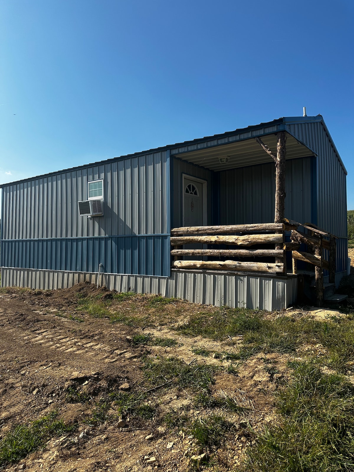 A cabin constructed with metal siding features a small porch supported by logs. A front door is visible, accompanied by a window and an air conditioning unit on the side. The surrounding area includes dirt paths and green grass under a clear blue sky.