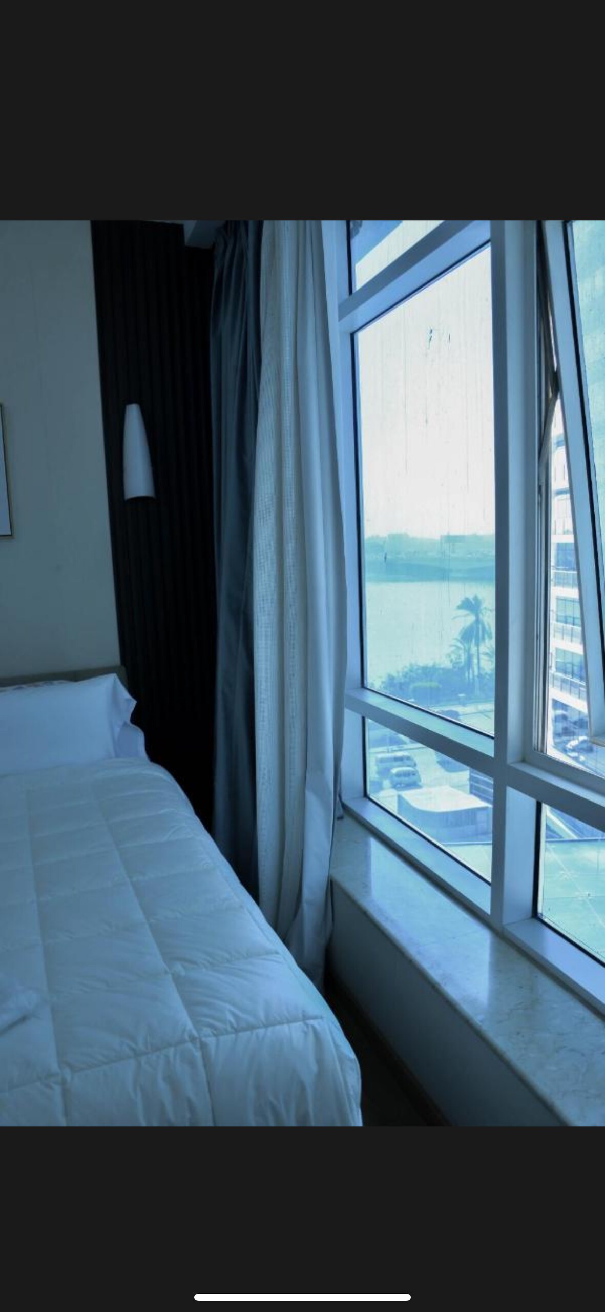 A bright and airy bedroom features a spacious bed dressed in white linens. Large windows allow natural light to fill the space, offering views of the surrounding area, including water and palm trees. Soft curtains frame the windows, enhancing the serene atmosphere.