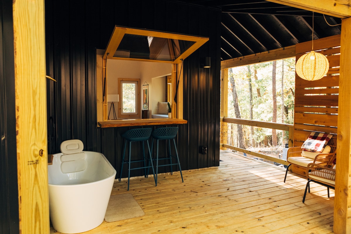 An inviting outdoor area features a modern soaking bathtub alongside a small bar with two stools. Natural wood flooring complements the black walls, while ample sunlight filters through the surrounding trees. A cozy lounge chair adds to the relaxed ambiance.