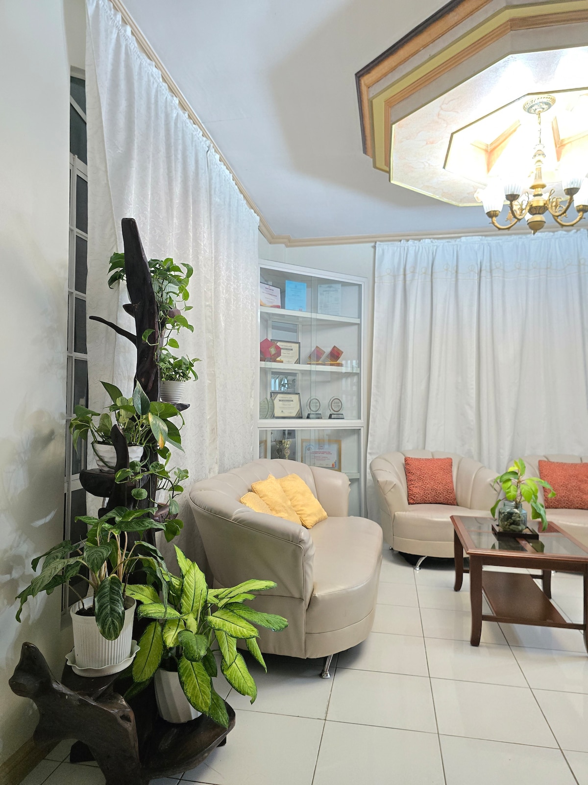 A welcoming living room area showcases a light-colored sofa and armchairs adorned with orange cushions. Potted plants add a touch of greenery. A wooden coffee table is centered in the space, complemented by a decorative shelving unit displaying awards, all under a softly illuminated chandelier.