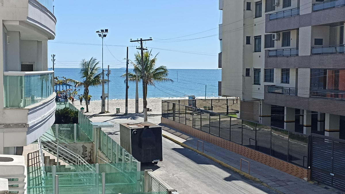 A clear view of the beachfront is presented, featuring palm trees and a sandy shore meeting the calm ocean waters. In the foreground, a street is visible, bordered by modern buildings and a gated entrance. The bright sky enhances the coastal atmosphere.