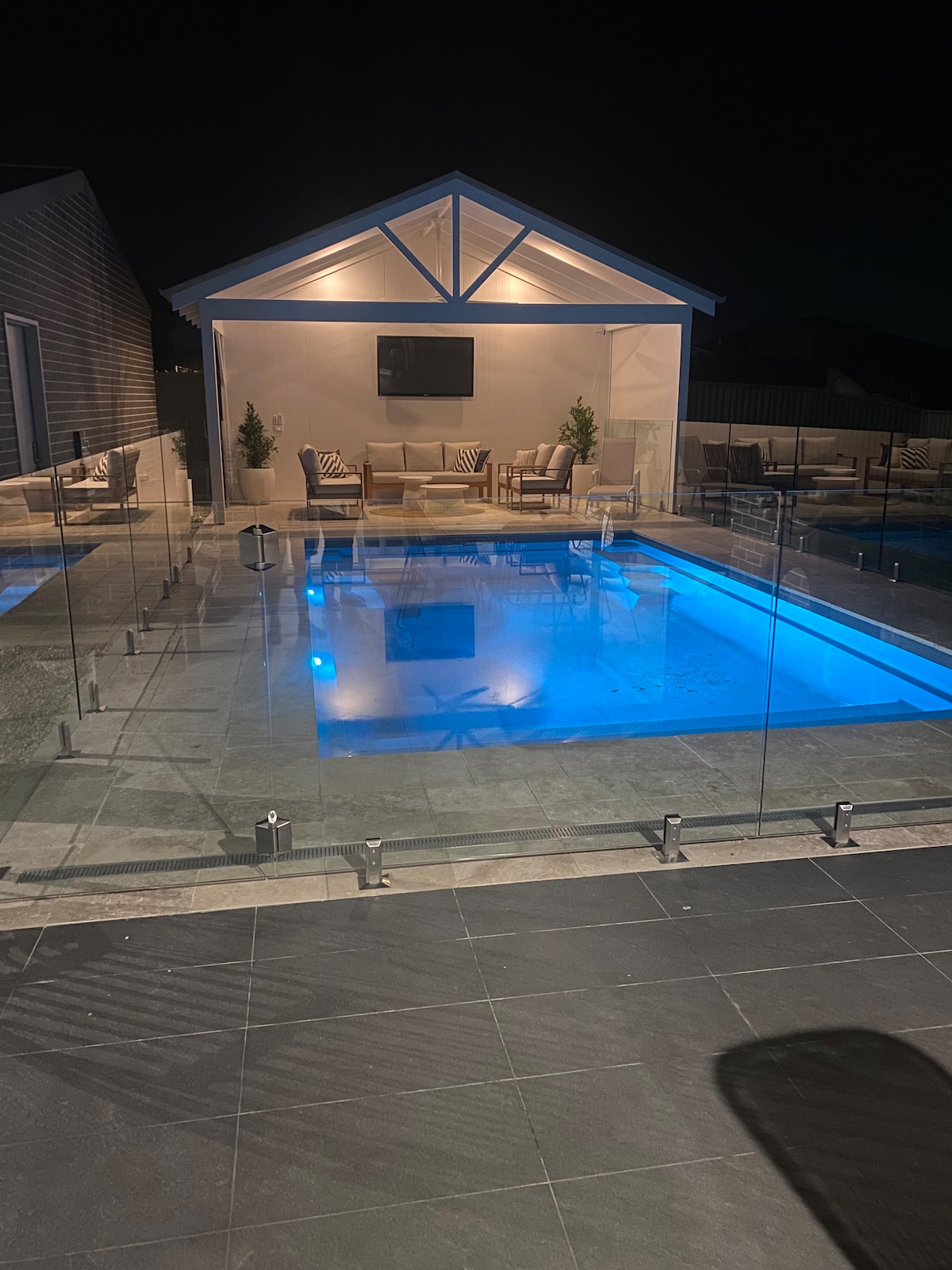 An illuminated pool area is showcased under a night sky, featuring a clear, blue water pool. A nearby lounge area includes plush seating and a wall-mounted television, creating a cozy atmosphere. Glass barriers provide safety while maintaining unobstructed views of the outdoor space.