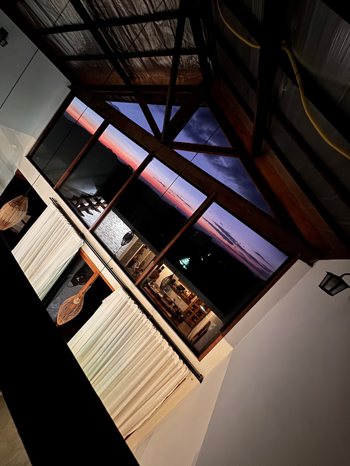 Large windows offer a panoramic view of the sunset, displaying vibrant colors reflected on the water. Wooden beams enhance the rustic charm of the space, while sheer curtains provide a soft barrier. The interior ambiance is accentuated by subtle lighting.