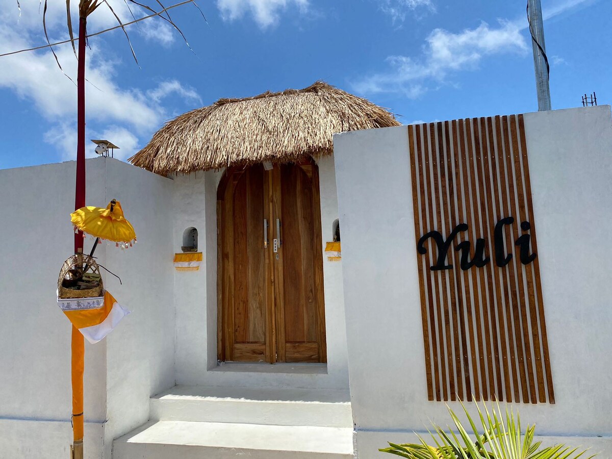 A welcoming entrance is framed by a thatched roof and wooden double doors. The white walls are complemented by wooden accents, with a decorative sign reading 'Puli.' A tropical plant is visible in the foreground, under a clear blue sky.