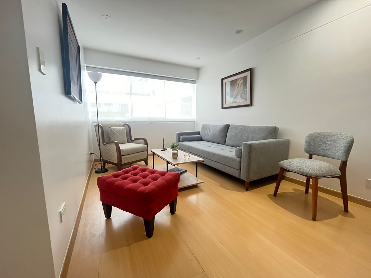 A comfortable living area is presented with a gray sofa, an accent chair, and a small red ottoman. Natural light fills the space through a large window, illuminating a wooden coffee table placed in the center. A floor lamp stands beside the sofa.