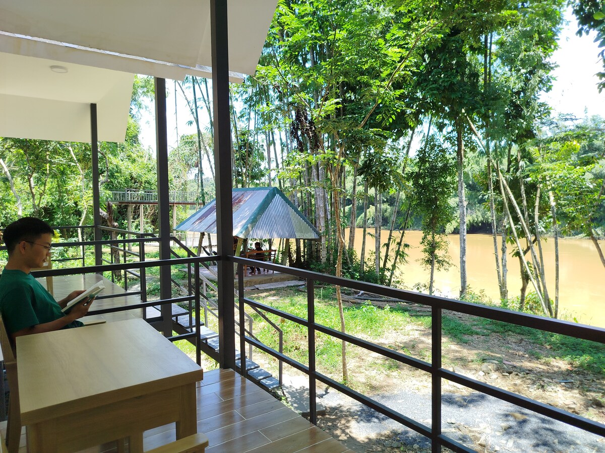 A shaded deck features wooden seating arrangements overlooking a river lined with lush greenery. Natural light filters through the trees, creating a serene atmosphere. The tranquil water reflects the surrounding vegetation, enhancing the peaceful setting ideal for relaxation.