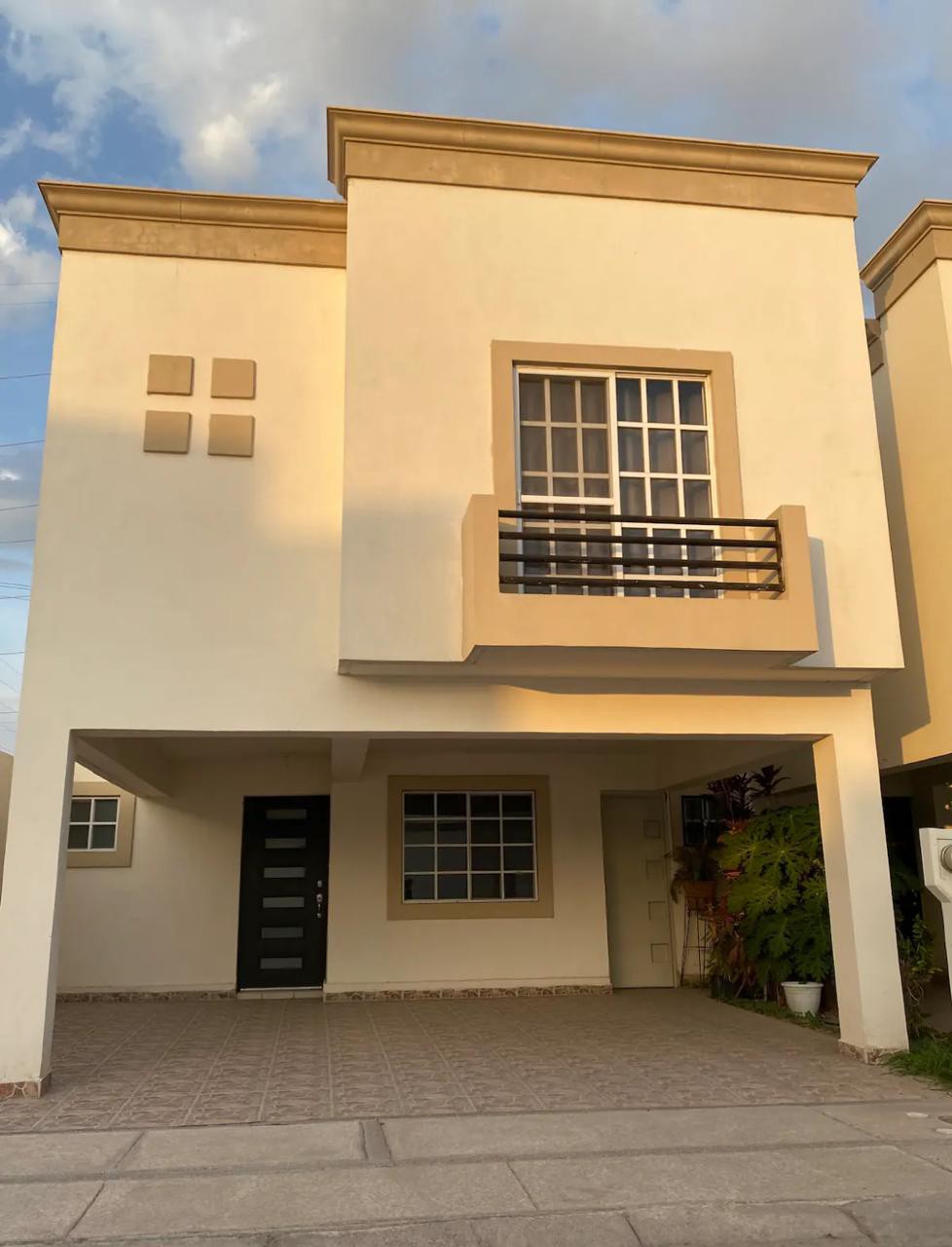 The exterior of a modern two-story home features a minimalist façade with a large window and a small balcony on the upper level. A spacious driveway is visible, leading to the entrance, bordered by potted plants. Soft evening light casts gentle shadows on the façade.