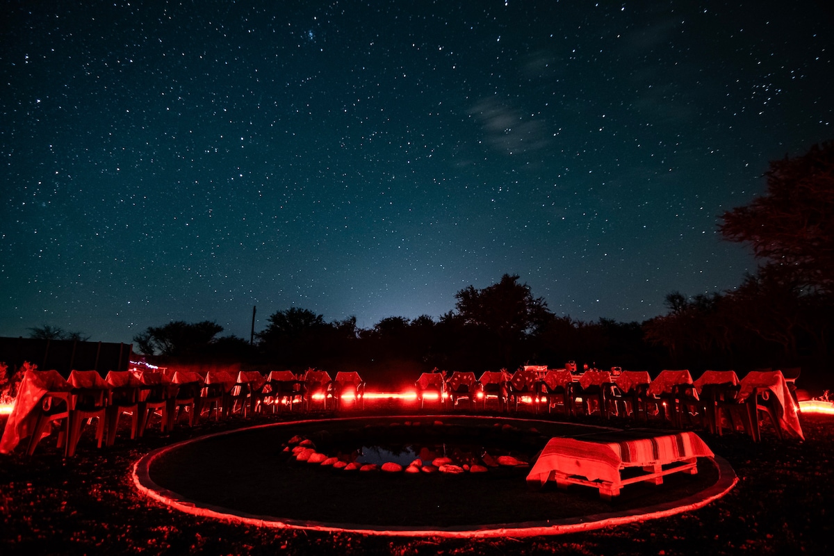 A circular area is illuminated by soft red lighting under a starry sky. Tables are arranged with white tablecloths, surrounded by chairs. A small pond is visible at the center, reflecting the starry night and creating a serene ambiance.