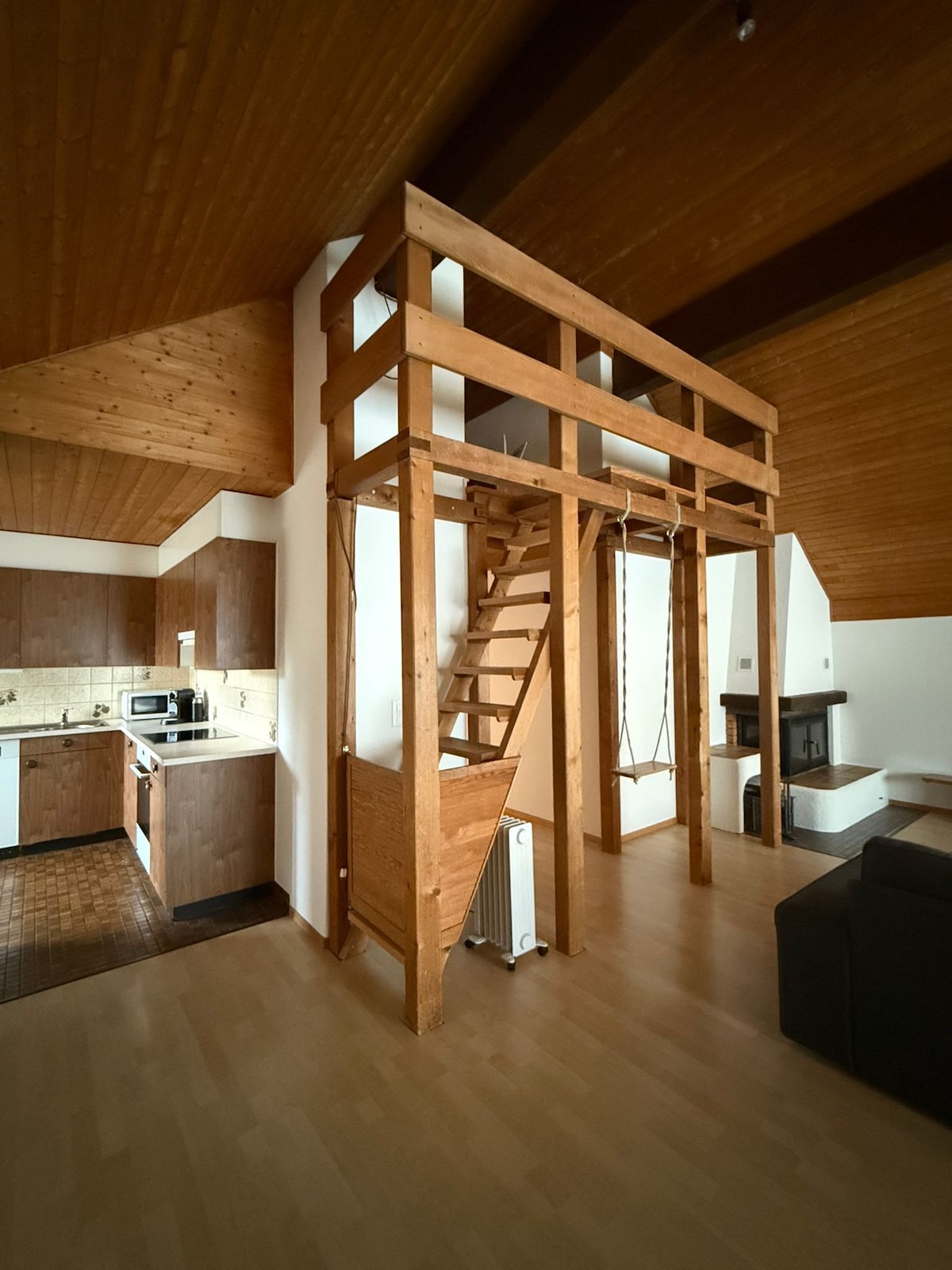 An open living space features a wooden loft structure accessed by stairs. The kitchen area includes modern appliances and cabinetry. Natural light fills the room through the high ceiling, accentuated by warm wood paneling. A cozy sitting area is visible in the lower section.