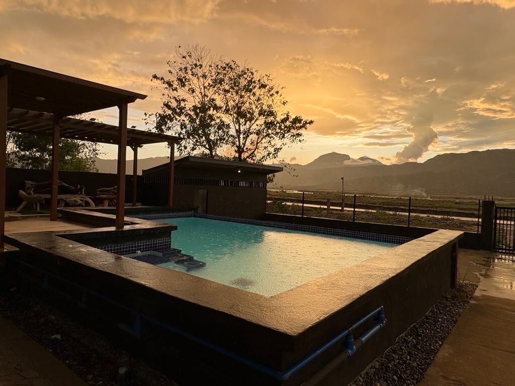 A serene pool area is framed by a wooden deck, with cozy seating options nearby. The sky is illuminated by a warm sunset, casting a golden glow over the view of distant mountains and lush trees, creating a peaceful outdoor setting.