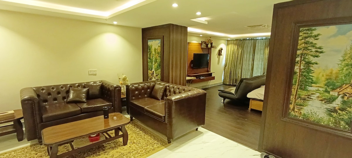 A living area features plush brown leather sofas arranged around a wooden coffee table. A flat-screen television is mounted on a decorative wall, complementing the artwork displayed in the room. Natural light filters in through the curtains, enhancing the comfortable environment.