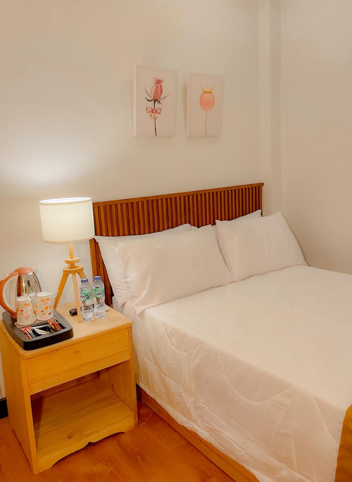 A cozy sleeping area is presented with a double-sized bed adorned with clean white sheets and four pillows. A bedside table features a lamp, a water kettle, and bottled water. Artwork is displayed on the wall above the bed, adding a touch of decor.