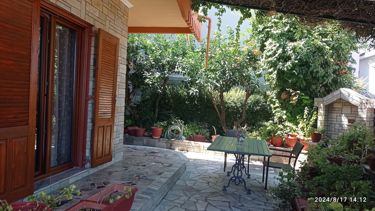 A stone pathway leads to an outdoor seating area, surrounded by lush greenery and potted plants. A table and chair set provide a space for relaxation, while the house features wooden shutters that enhance the natural light filtering in from the garden.
