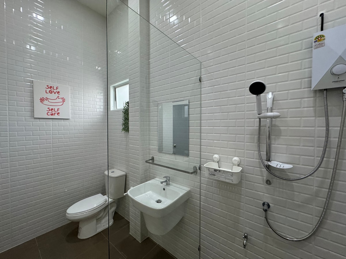A well-lit bathroom features modern fixtures, including a glass-enclosed shower and a wall-mounted showerhead. The minimalist design is complemented by white tiled walls and a decorative plant. A motivational wall art piece is displayed above the toilet.