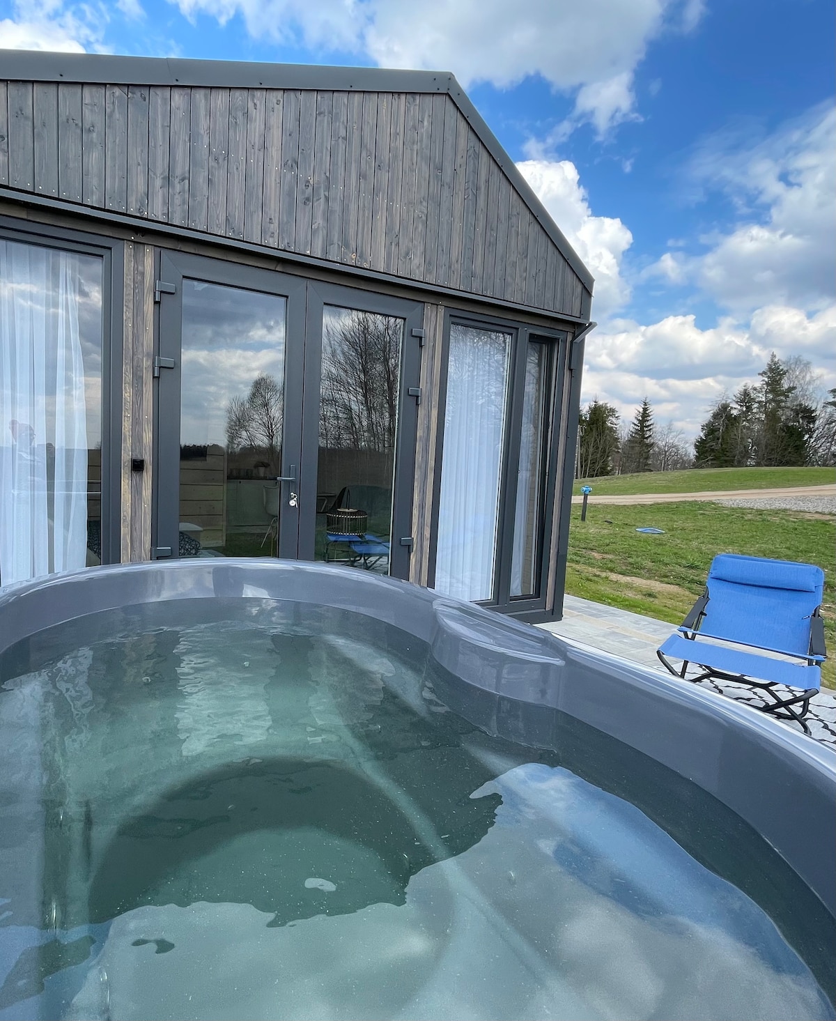 A spacious outdoor terrace is highlighted by a natural wooden hot tub, featuring clear water that reflects the sky. Modern glass doors leading into the interior provide an inviting connection to the surroundings. A single blue chair is positioned nearby, complementing the tranquil setting.