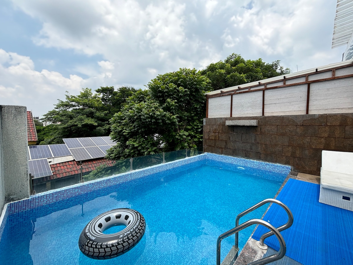 A rooftop swimming pool features clear blue water surrounded by stone walls and a glass railing. A circular float is present in the pool, with greenery and solar panels visible in the background. Sunlight filters through scattered clouds overhead.