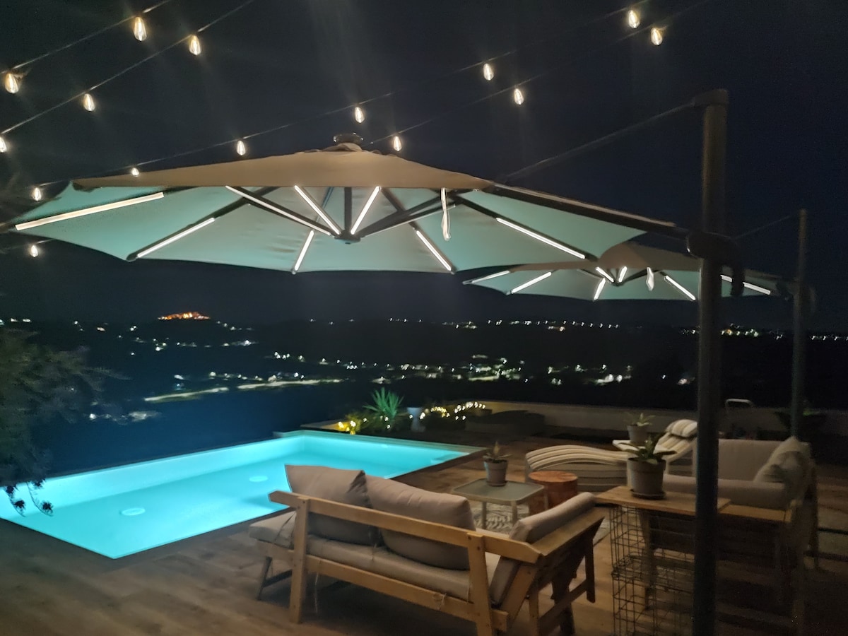 An outdoor patio area features a softly illuminated infinity pool and comfortable seating under spacious umbrellas. String lights add a warm glow to the space, with views of the surrounding landscape visible in the background at night.