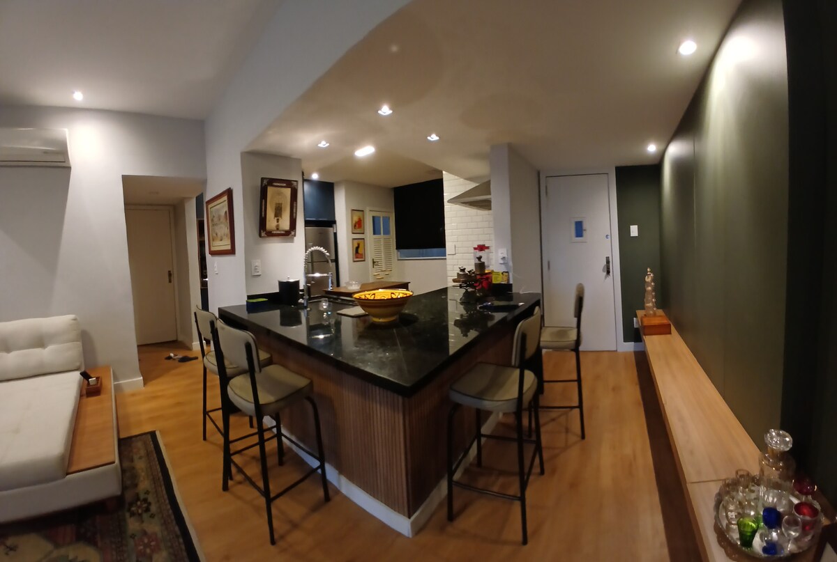 An open kitchen area features a large dark countertop with seating for four. Light fixtures highlight the space, complemented by a bowl of fruit. A dining area is visible, accompanied by a wooden bench and decorative elements on a shelf against the wall.