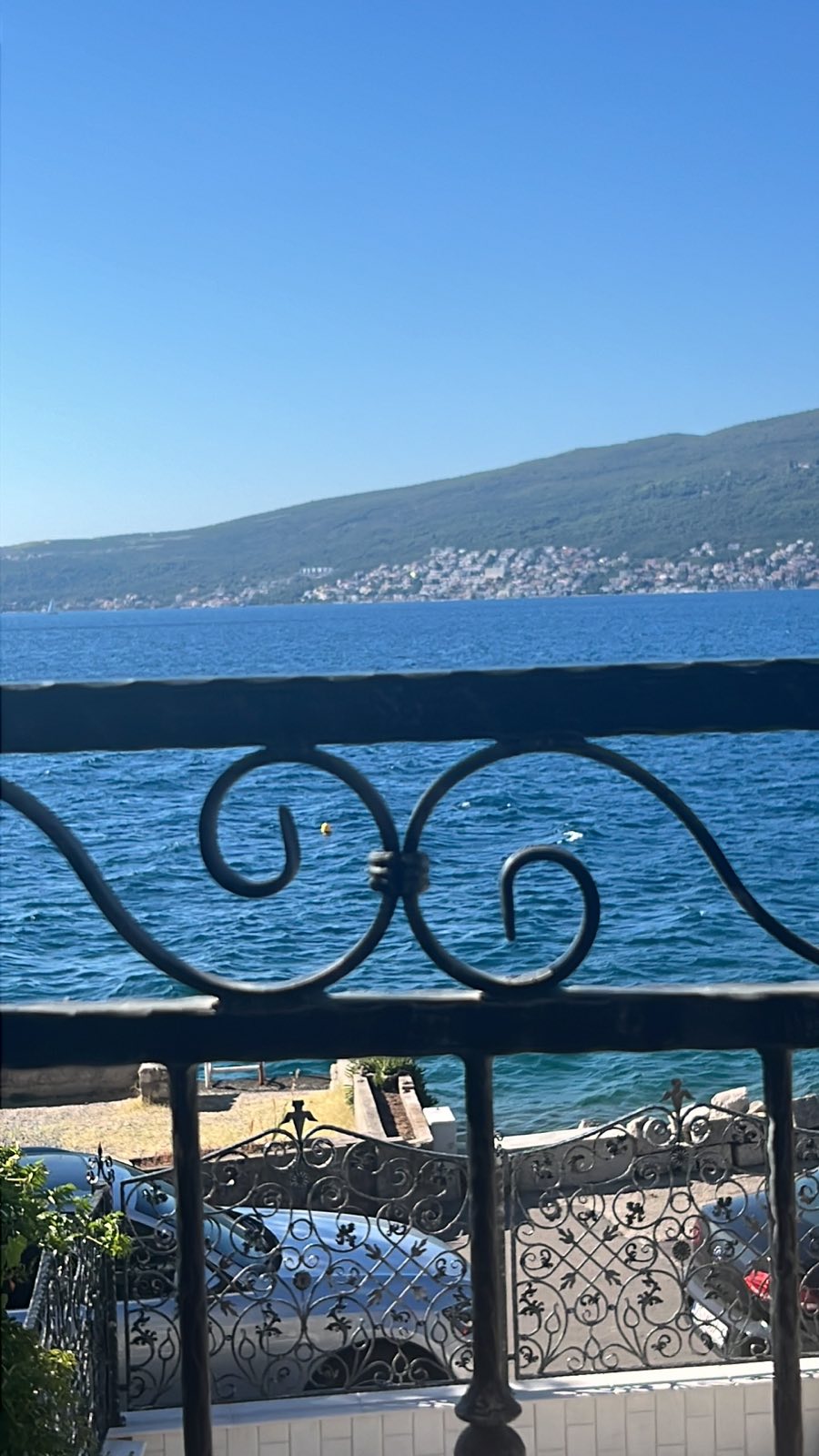A balcony view overlooks the serene Adriatic Sea, framed by an ornate iron railing. The shimmering water reflects sunlight, and distant hills rise gently in the background, creating a sense of tranquility. The scene captures a calm, inviting atmosphere by the seaside.