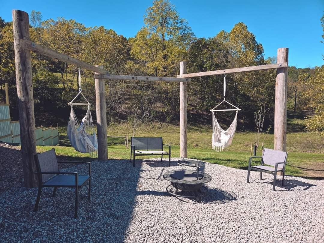 A serene outdoor space features a cozy seating arrangement with two chairs and a fire pit, surrounded by gravel. Two hanging hammocks provide additional relaxation options, framed by wooden beams, with greenery and rolling hills visible in the background.