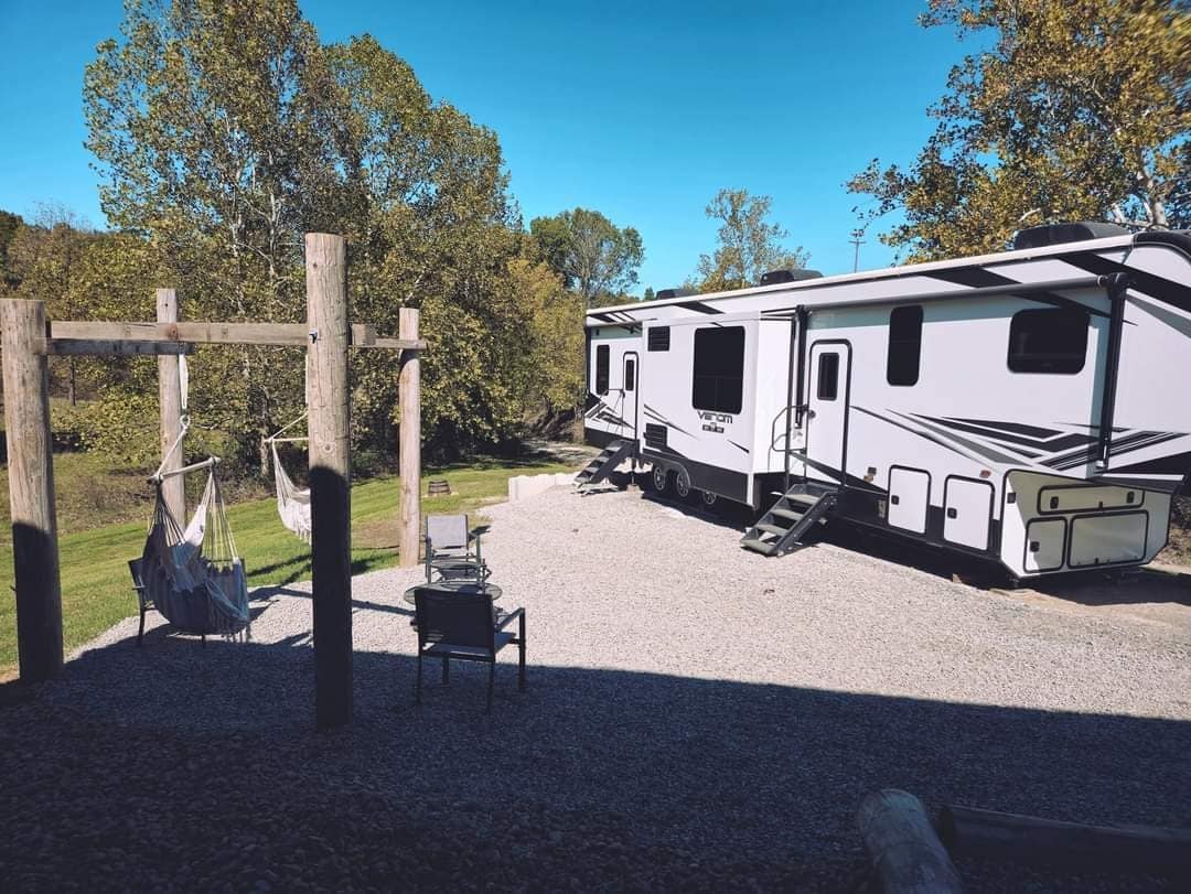 A fifth wheel RV is situated on a gravel area under a clear blue sky. Nearby, a wooden swing set and a hammock invite relaxation. The surrounding landscape features lush trees and green grass, offering a serene outdoor environment.