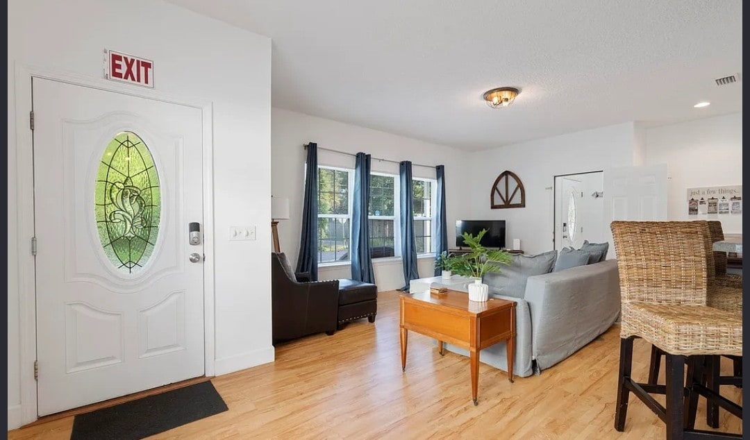 A welcoming entryway features a decorative glass door, leading to a spacious living area with large windows allowing natural light. Comfortable seating includes a sleek leather armchair and a soft sectional couch, complemented by a wooden coffee table and an inviting dining space with woven chairs.