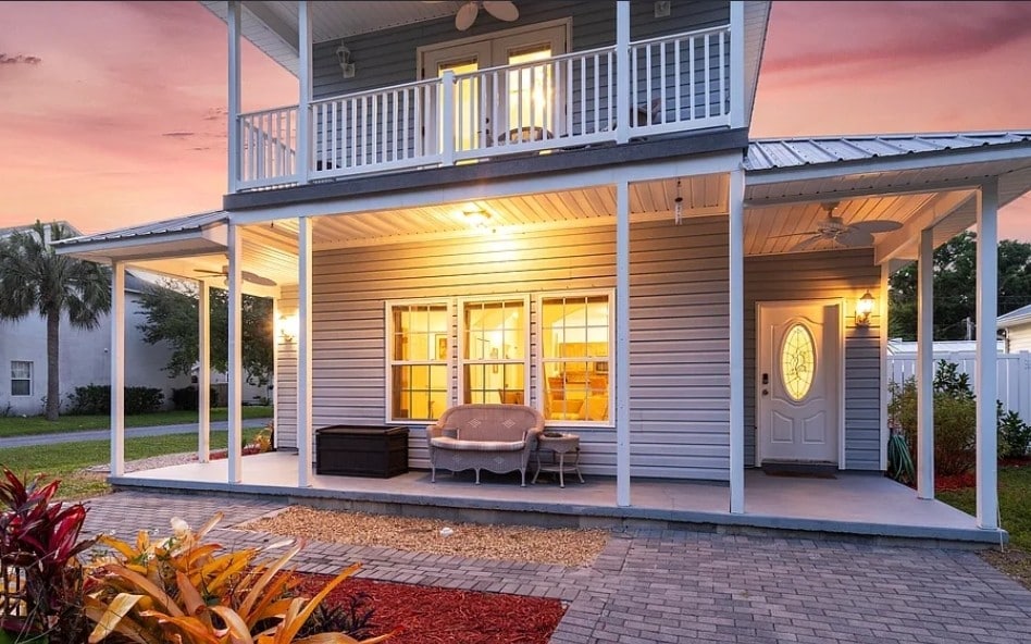 A two-story home features a spacious front porch with comfortable seating and large windows that illuminate the interior. Lush landscaping and paving stones lead to the entrance. Vibrant plants complement the exterior, while a serene sunset provides a warm glow.