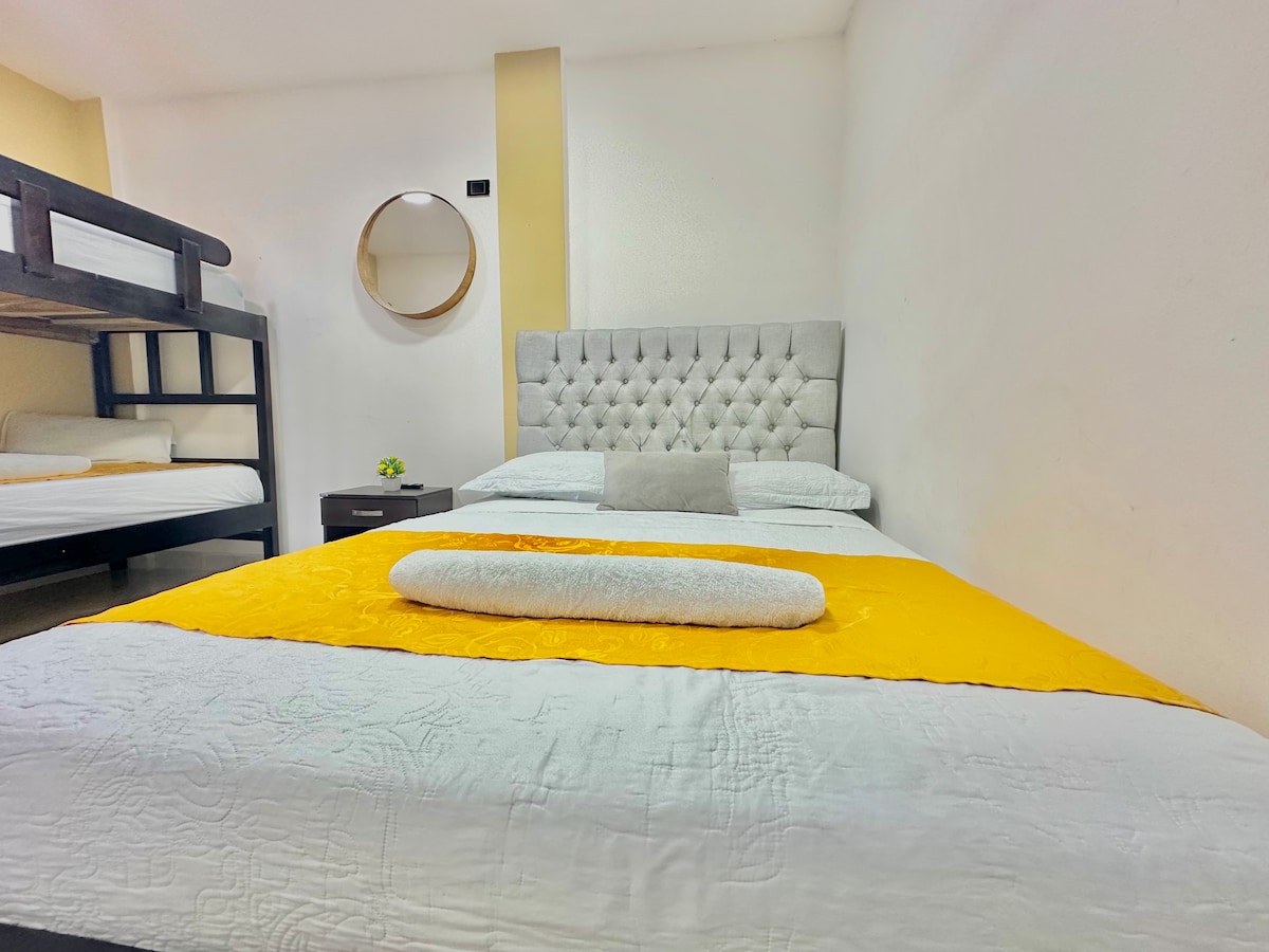 A cozy sleeping area features a spacious bed with a tufted gray headboard and a yellow accent blanket. A circular mirror is mounted on the wall, and a small decorative plant rests on a dark nightstand. A bunk bed with a simple design is visible in the background.