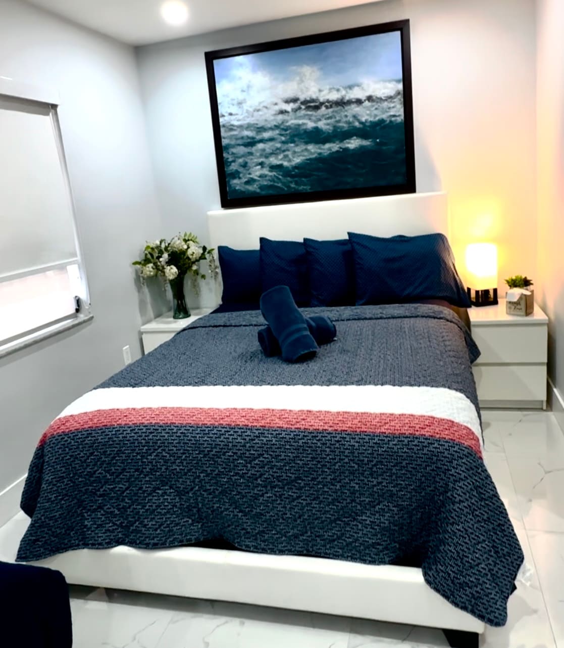 A cozy bedroom features a spacious bed dressed in a patterned navy-blue coverlet with complementary pillows arranged at the headboard. A piece of abstract art hangs above the bed, and a bedside table with a lamp and small plant is positioned next to the bed. Natural light enters through a window with shades.