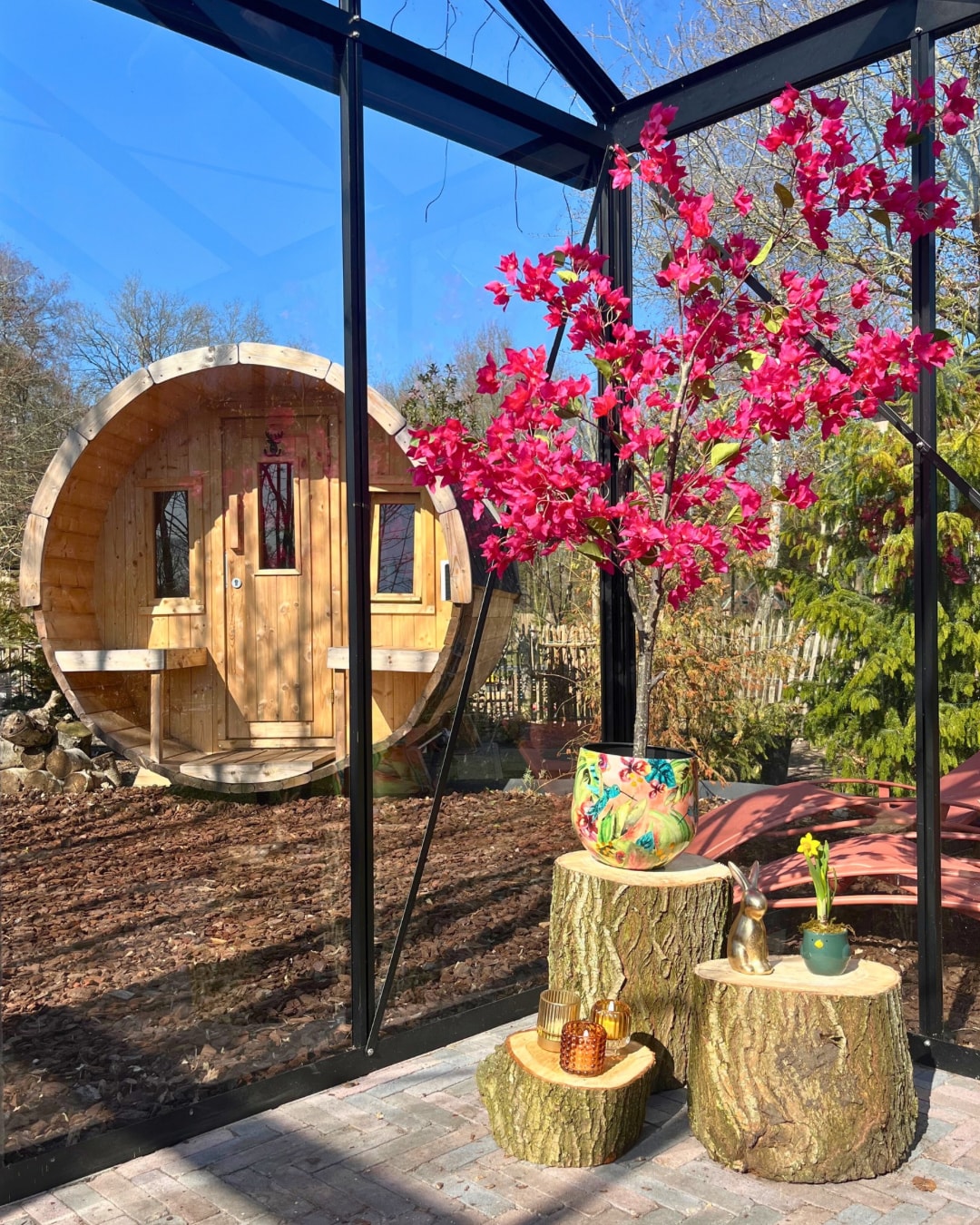 A wooden barrel-shaped accommodation is visible through a glass structure, surrounded by nature. Vibrant pink flowers in a decorative pot are placed on a tree stump, enhancing the natural ambiance. The outdoor setting includes a rustic feel with greenery and a calm atmosphere.