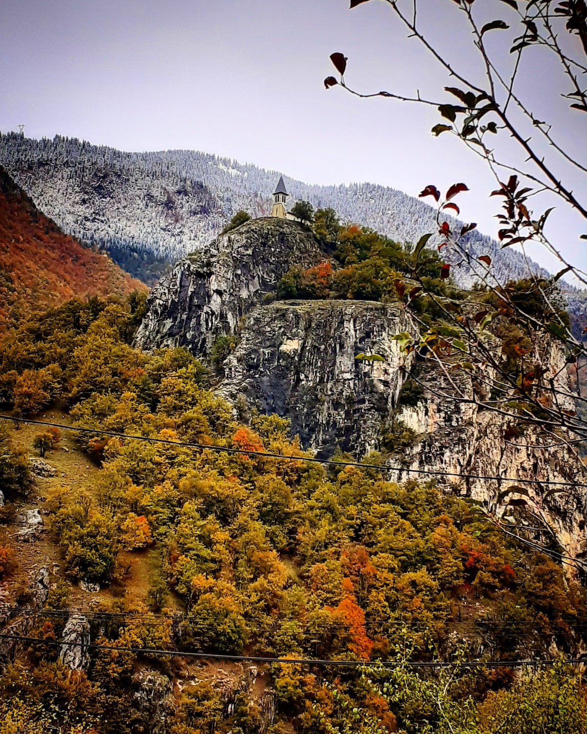 A mountain landscape is depicted, featuring a rocky outcrop topped with a small chapel. The surrounding mountains display a mix of green and orange foliage, indicating the season. Snow-capped elevations rise in the background, contrasting with the vibrant autumn colors below.