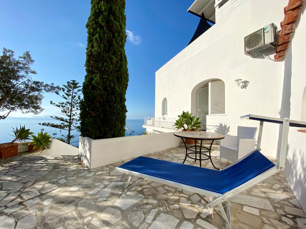 An outdoor terrace is presented, featuring a blue lounge chair and a small round table. Lush greenery and well-maintained stone flooring add to the inviting space. The backdrop reveals ocean views under a clear blue sky, creating a serene atmosphere.