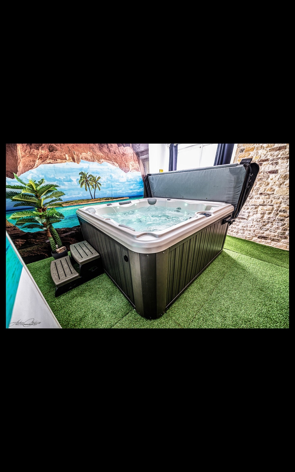 A spacious hot tub is situated in a room with attractive stone walls. The tub is equipped with steps for easy access, and a mural depicting a tropical landscape adds to the ambiance. A green carpet enhances the relaxation setting.