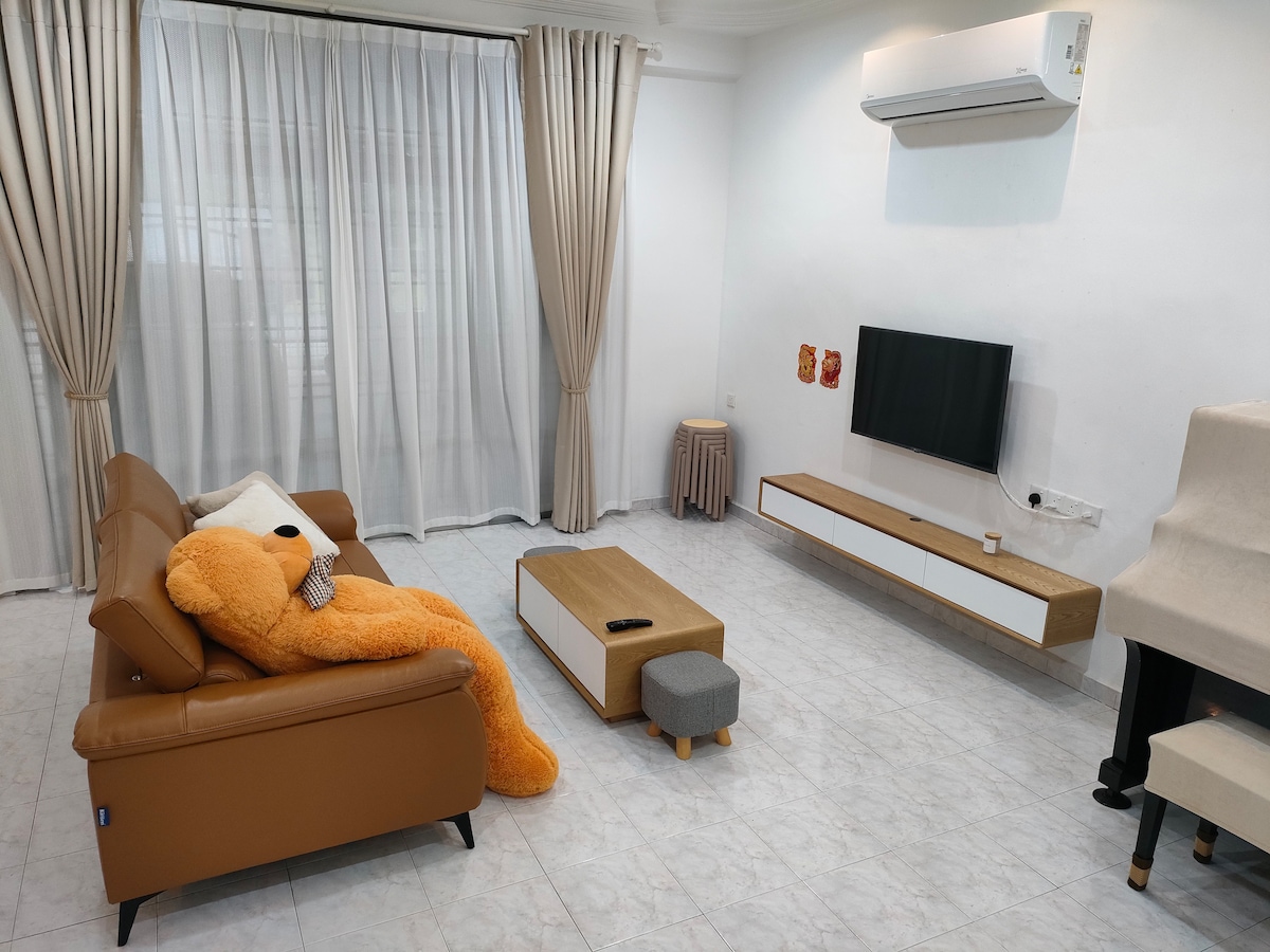 A spacious living area is presented with a tan sofa featuring a plush orange teddy bear. A low coffee table sits in front of the sofa, with a small footrest nearby. A wall-mounted television is positioned opposite the seating, with sheer curtains allowing light to enter.