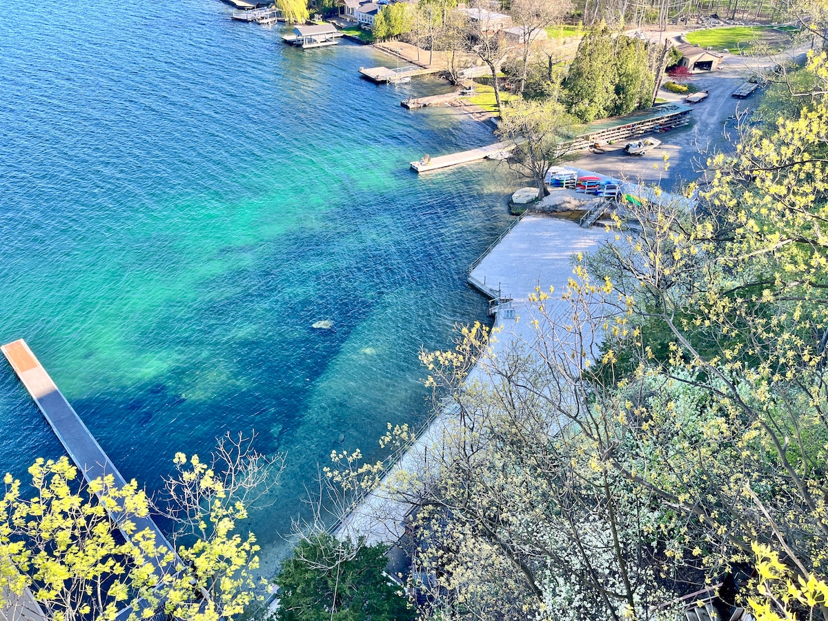 A serene view of Canandaigua Lake is captured from above, showcasing clear blue waters and a mix of sandy and grassy shoreline. Docks are visible along the water's edge, with trees surrounding the area, creating a tranquil setting for outdoor activities.