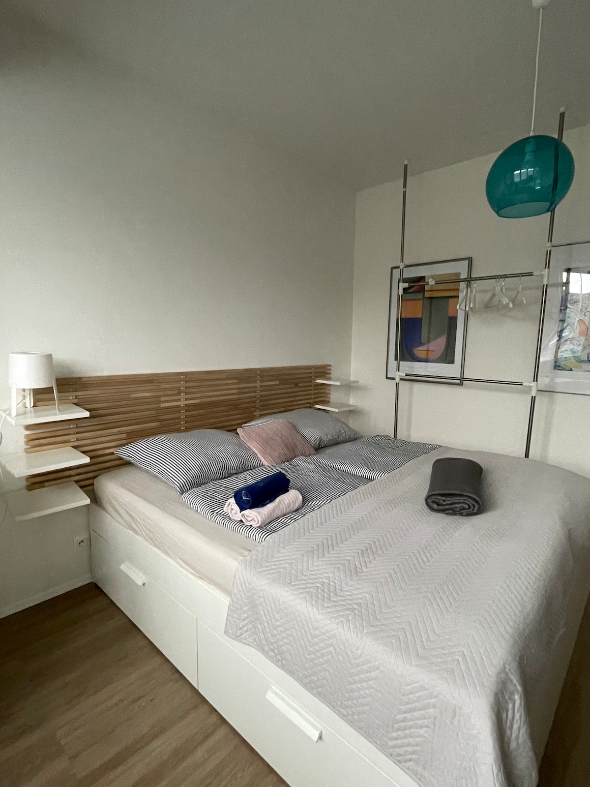 A serene sleeping area features a spacious bed with muted bedding, complemented by a wooden headboard. Soft lighting is provided by a small lamp and a hanging glass pendant. The room is decorated with minimalistic shelves and wall art, enhancing a calming atmosphere.