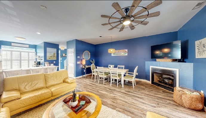 A spacious living area features a light-colored sofa and armchair, with a glass coffee table at the center. A large dining table is arranged near the kitchen, and a flat-screen television is mounted on the blue wall above an electric fireplace, providing a cozy ambiance.