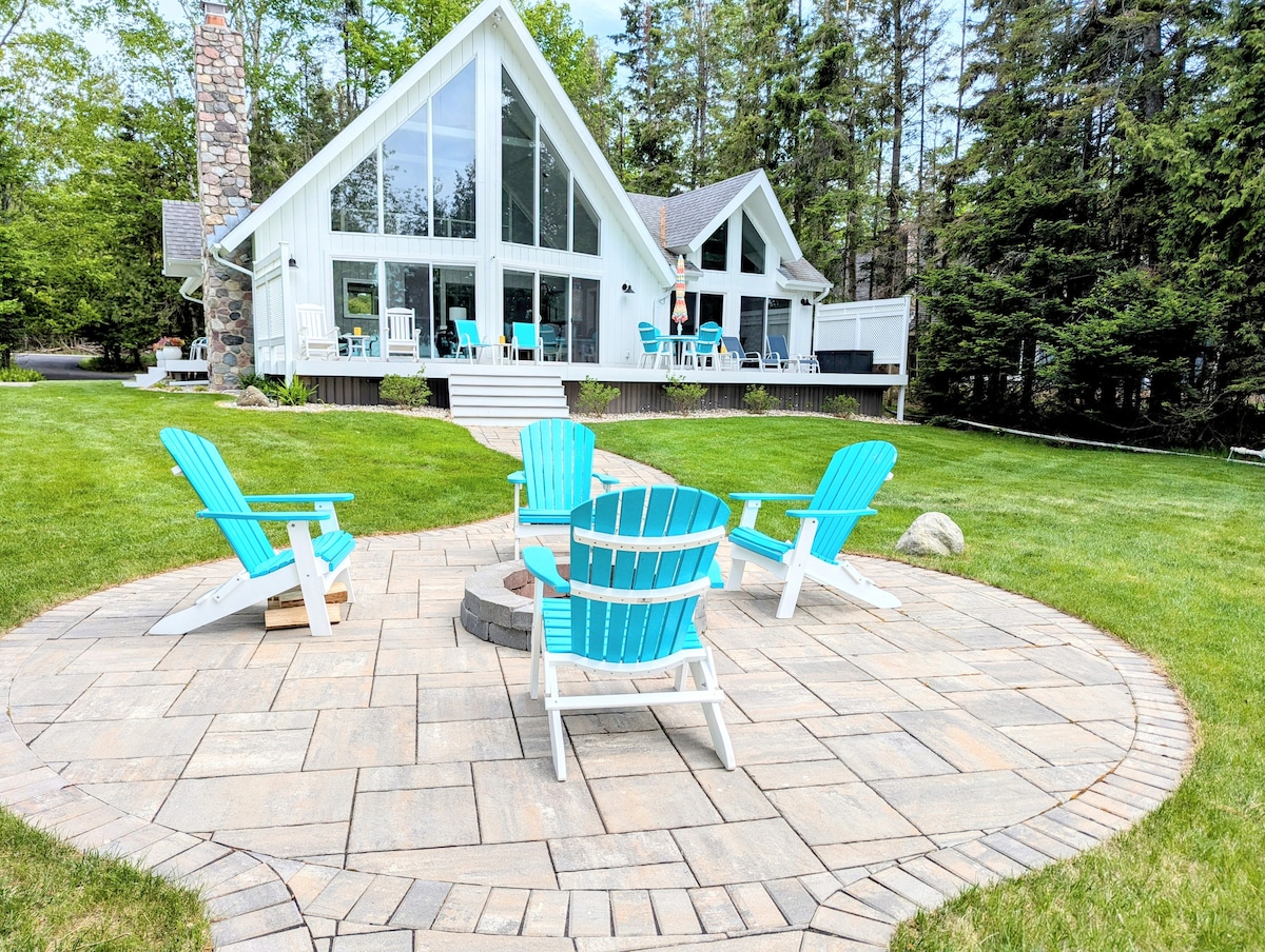 A circular stone patio features four colorful adirondack chairs arranged around a fire pit. The cottage's large windows reflect a natural setting, framed by lush green grass and trees. Outdoor seating and a white deck can be seen in the background, providing a relaxing atmosphere.