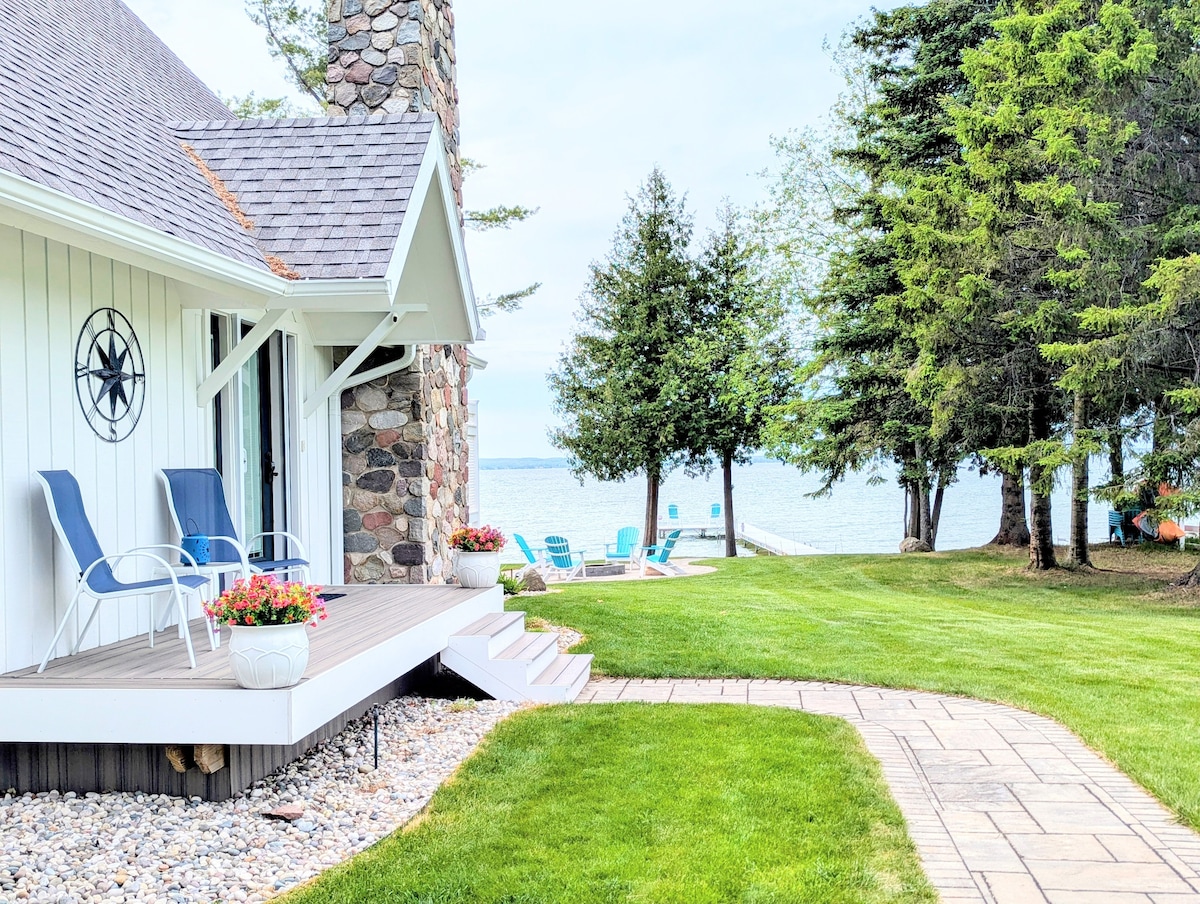 The exterior of the cottage features a stone chimney and a welcoming porch with two chairs. Lush green grass surrounds a paved pathway leading towards Mullett Lake. Colorful planters add vibrancy, while glimpses of the dock and seating are visible in the background, framed by tall trees.